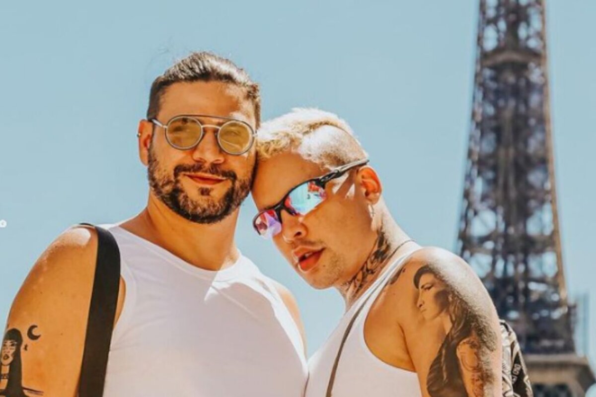 Gloria Groove e o marido, ambos de óculos e camiseta branca, com a Torre Eiffel ao fundo