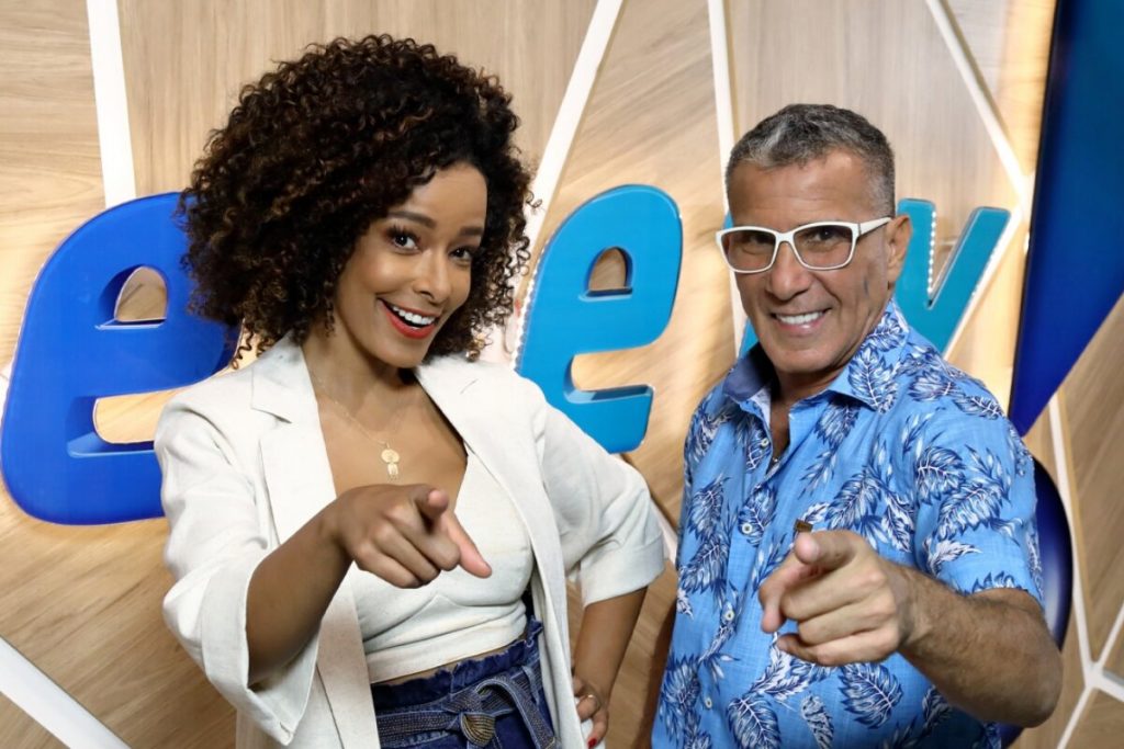 Alline Prado de branco e Eri Johnson de azul, diante da logo da Rede TV, ambos apontado para a frente e sorrindo