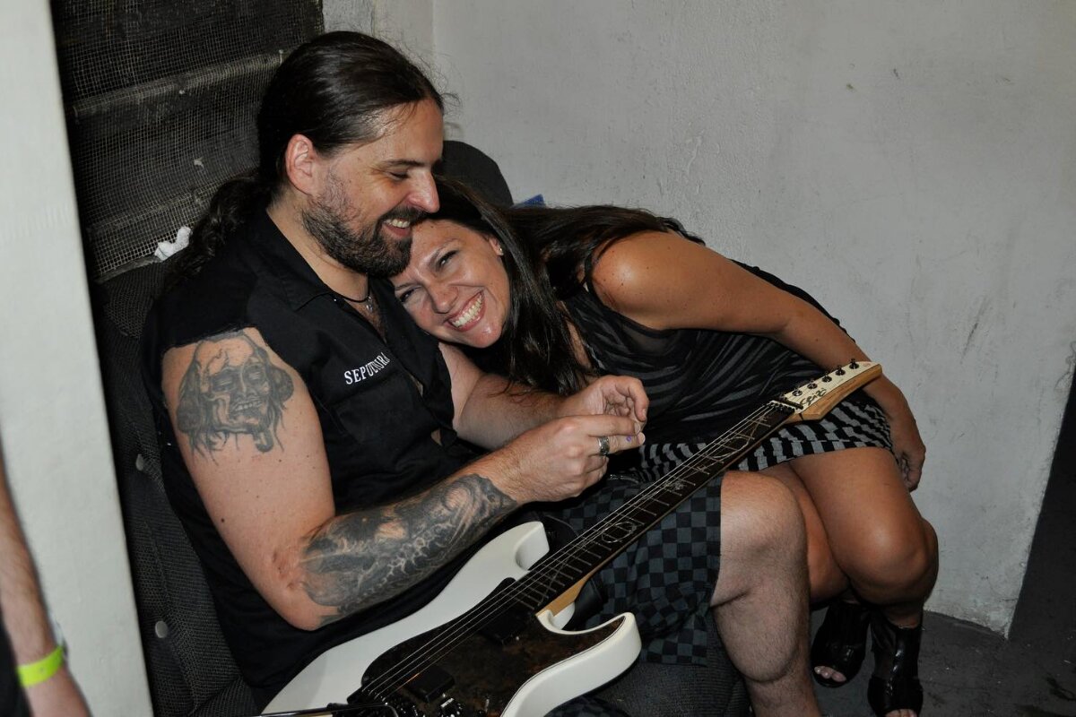 Andreas Kisser segurando uma guitarra, sentado no chão com a mulher, Patrícia, deitada no colo dele