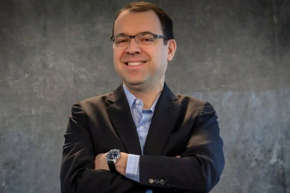 Mário Teixeira de terno preto, camisa social azul, de braços cruzados, de óculos, sorrindo