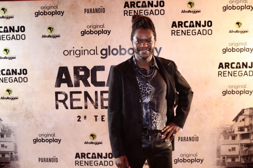 Bukassa Kabengele todo de preto, sorrindo, posando no backdrop de Arcanjo Renegado