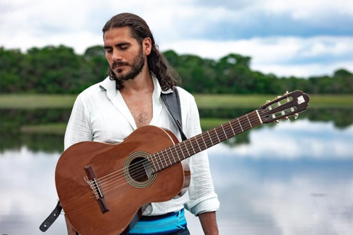 Gabriel Sater, olhando de lado, em frente ao rio, com viola pendurada no ombro