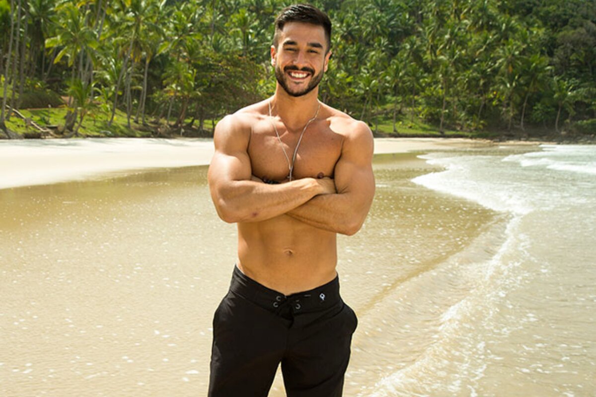 Igor Freitas sem camisa, de raços cruzados, bermuda preta, numa praia