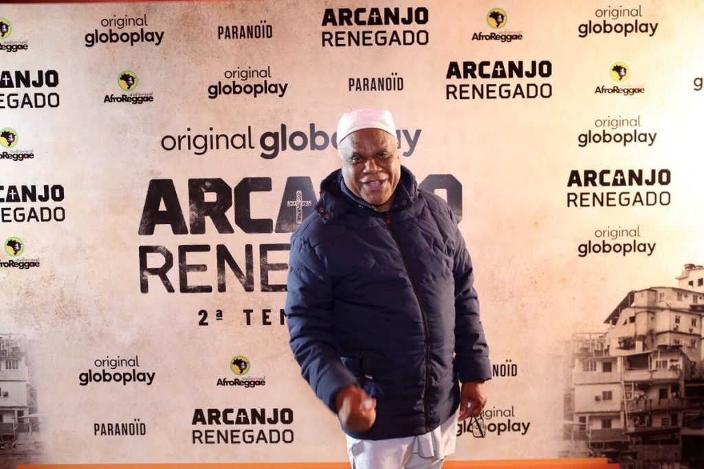 Ivanir dos Santos de jaqueta azul escura, calça branca e gorro branco, posado no backdrop de Arcanjo Renegado