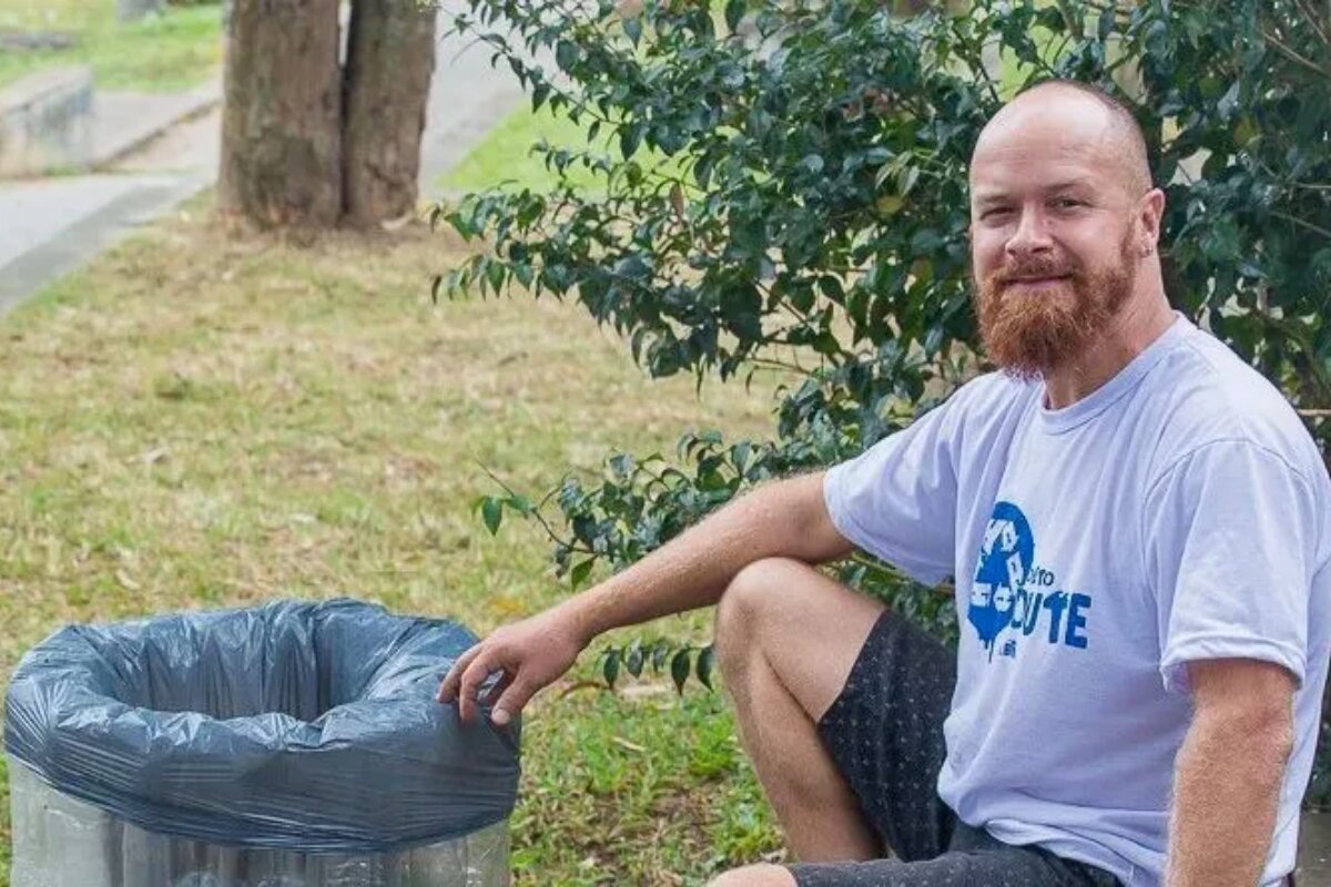 Ambientalista Ferrugem, agachado diante de uma lata de lixo, de camisa branca e bermuda preta