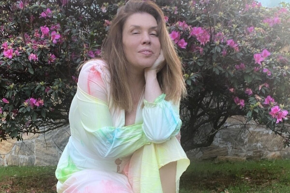 Simone de vestido tie-dye, ajoelhada no jardim, com a mão no pescoço