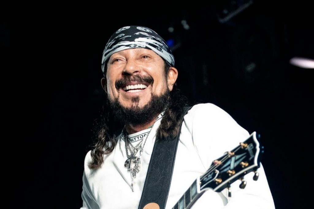 Bell Marques com a bandana na cabeça, camisa branca, tocando guitarra e sorrindo