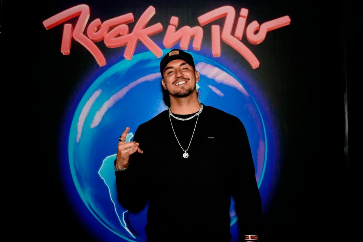 Gabriel Medina de boné preto, casaco preto, fazendo sinal de hang loose, sorrindo, diante doa logo do Rock in rio