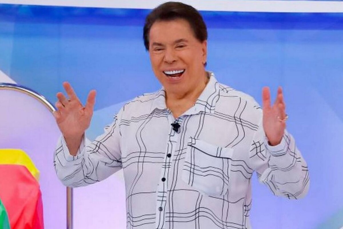 Silvio Santos de camisa social branca de listras finas pretas, de braços abertos, sorrindo