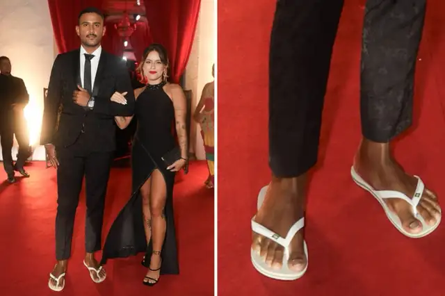 Lucas Pizane de terno preto e chinelo branco, de braços ados com a namorada, de vestido preto