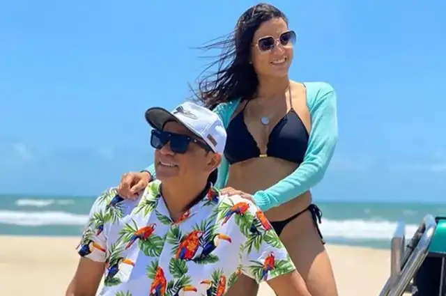 Frank Aguiar de boné branco, camisa florida, com Carol Santos, de biquíni preto e camisa de manga verde, na praia